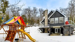 A wooden play structure with a slide and a colorful canopy, a stone chimney, and a house with a balcony and a covered porch. - Stillwater Lakehouse • 20 Acre Lakefront • Sandy Beach • Dock • Firepit! (Port Carling)