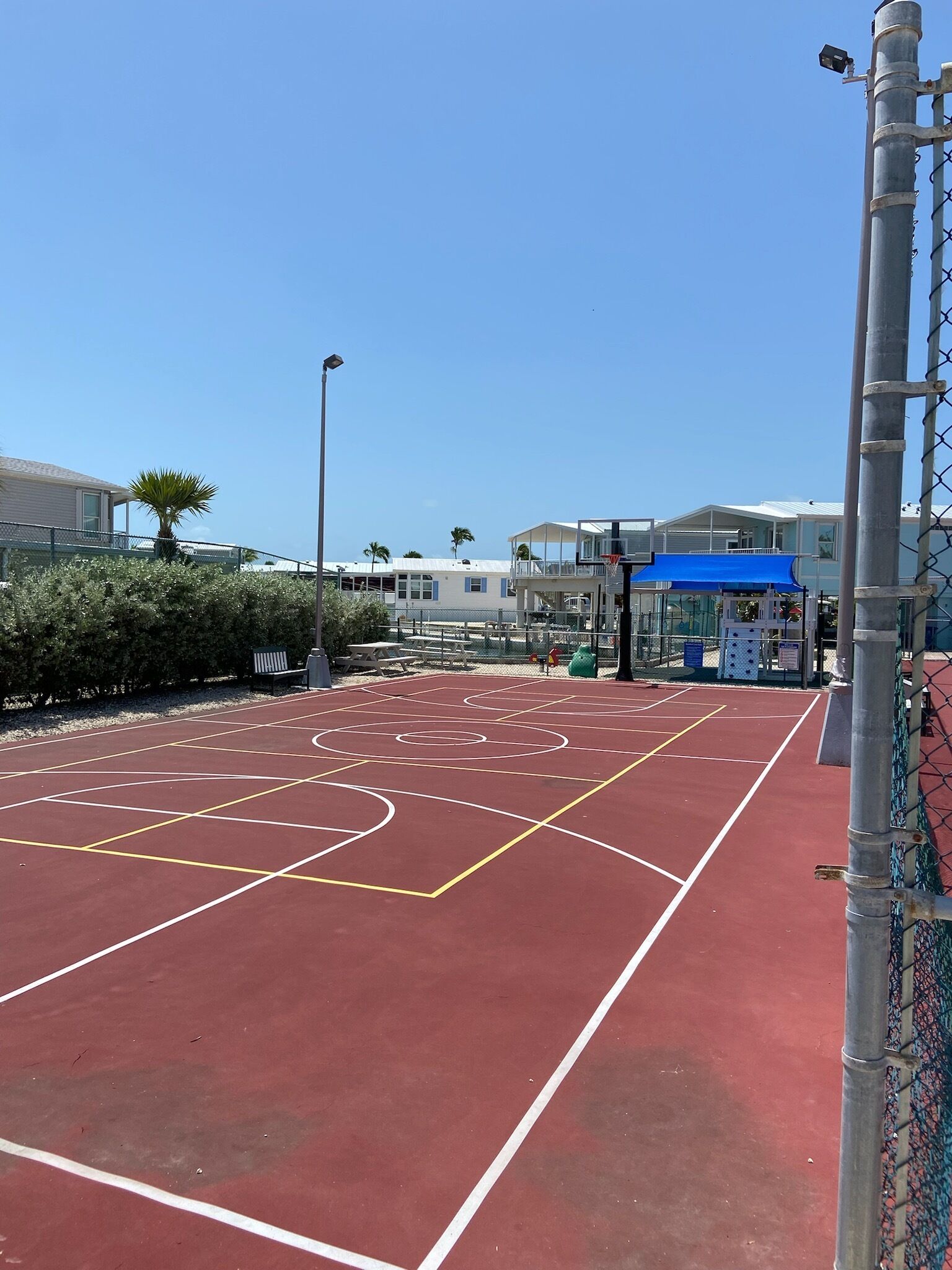 An outdoor basketball court with a red surface, surrounded by a chain-link fence, and a blue canopy structure in the background.