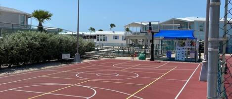 An outdoor basketball court with a red surface, surrounded by a chain-link fence, and a blue canopy structure in the background.
