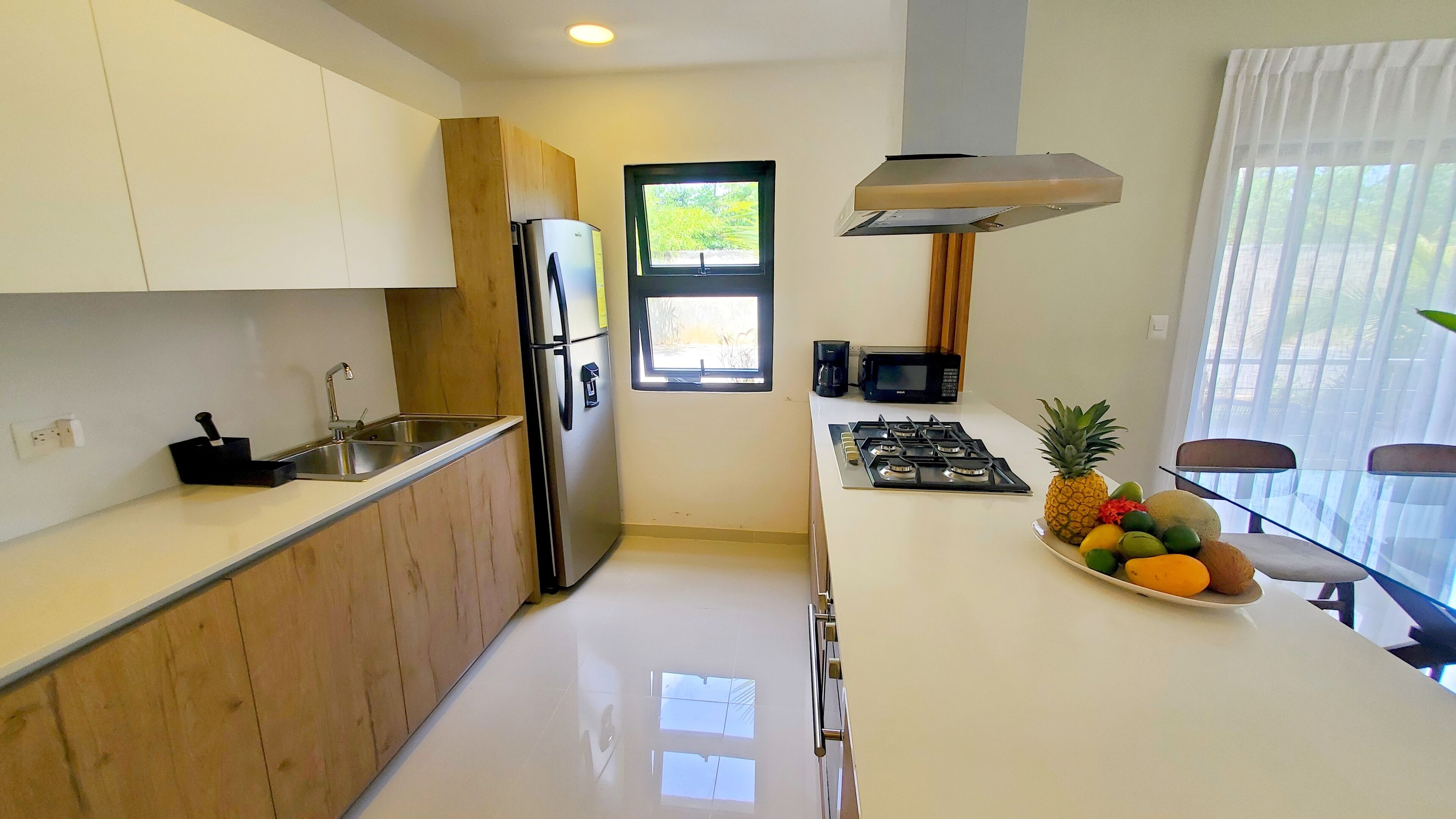 A modern kitchen with white cabinets, a stainless steel refrigerator, a microwave, and a gas stove. There's a window providing natural light and a fruit bowl on the counter.