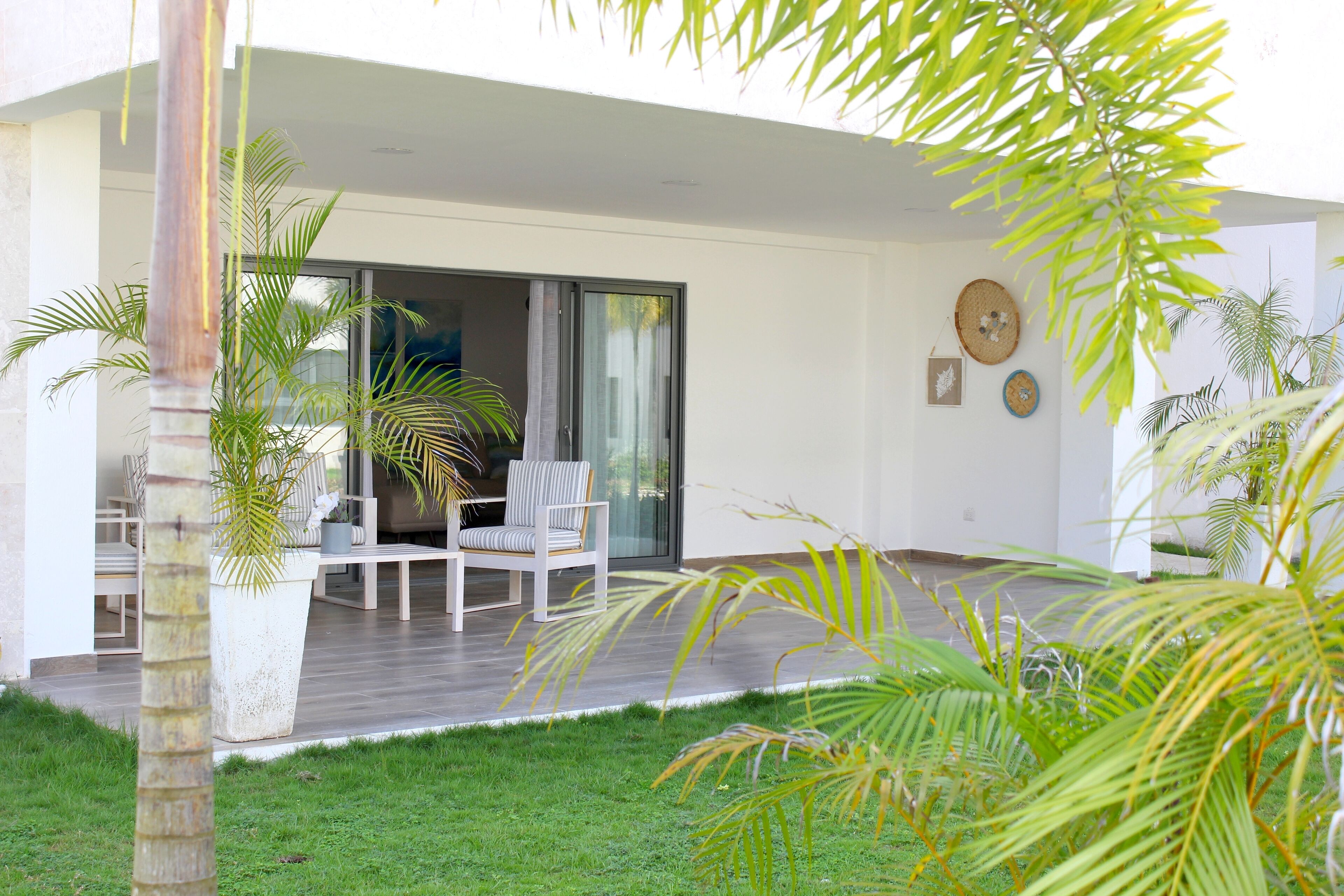 Un patio moderno al aire libre con un ventanal de vidrio que da a una sala de estar, rodeado de abundante vegetación y una terraza de madera.
