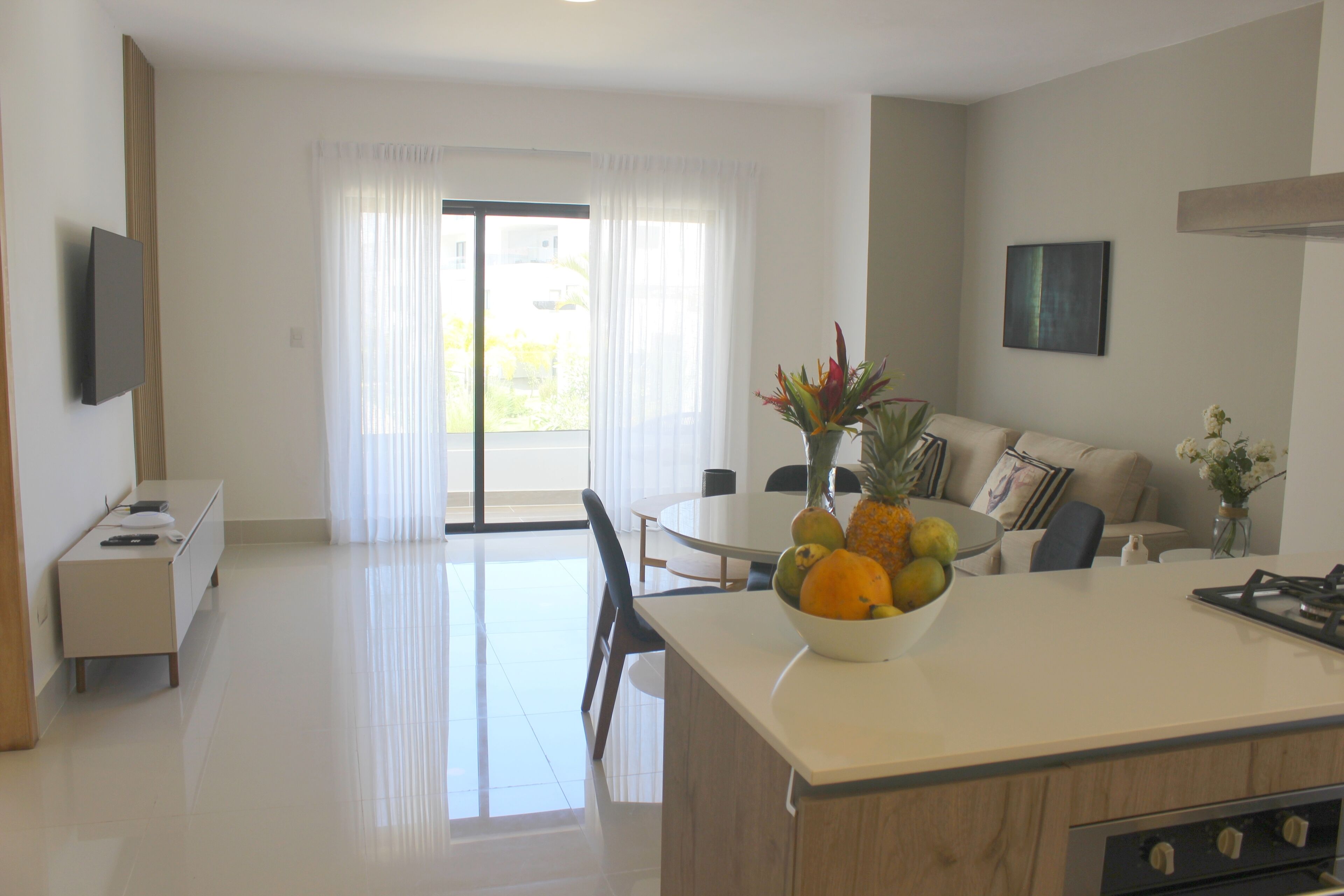 A modern kitchen with a central island, a dining area with a round table, and a living space with a sofa and a TV mounted on the wall.