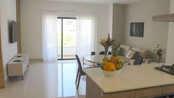 A modern kitchen with a central island, a dining area with a round table, and a living space with a sofa and a TV mounted on the wall.