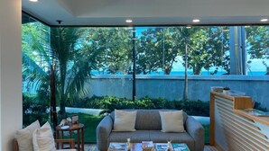A modern living room with a sofa, armchair, and coffee table, large windows with a view of greenery and a building. - Amar'e Aparthotel & Spa (Las Terrenas)
