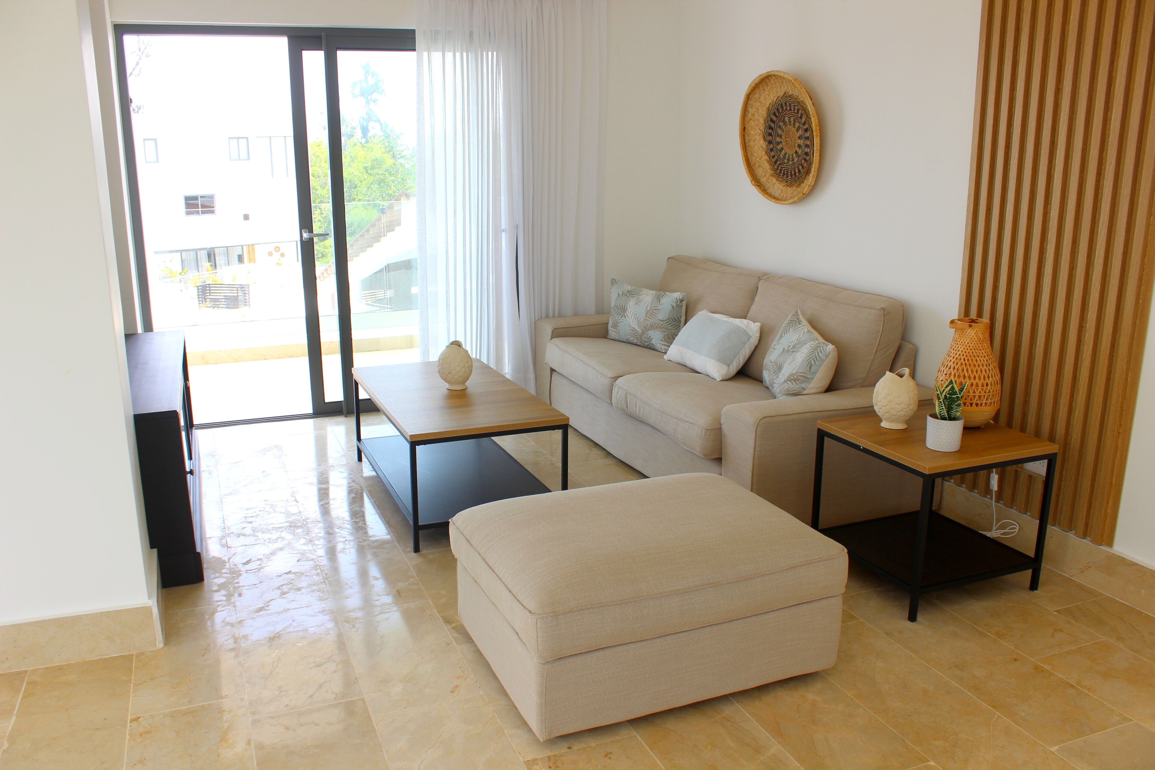 A modern living room with a sofa, ottoman, and coffee table, a sliding glass door leading to an outdoor area, and a decorative wall piece.