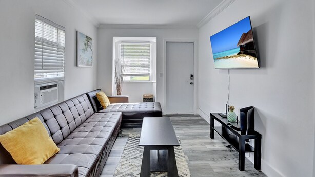Un salon moderne avec un canapé d’angle, une table basse, un téléviseur à écran plat fixé au mur et une grande fenêtre avec des stores.