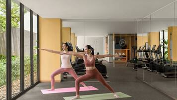 Two people doing yoga on mats in a room with large windows, exercise equipment, and a view of greenery.