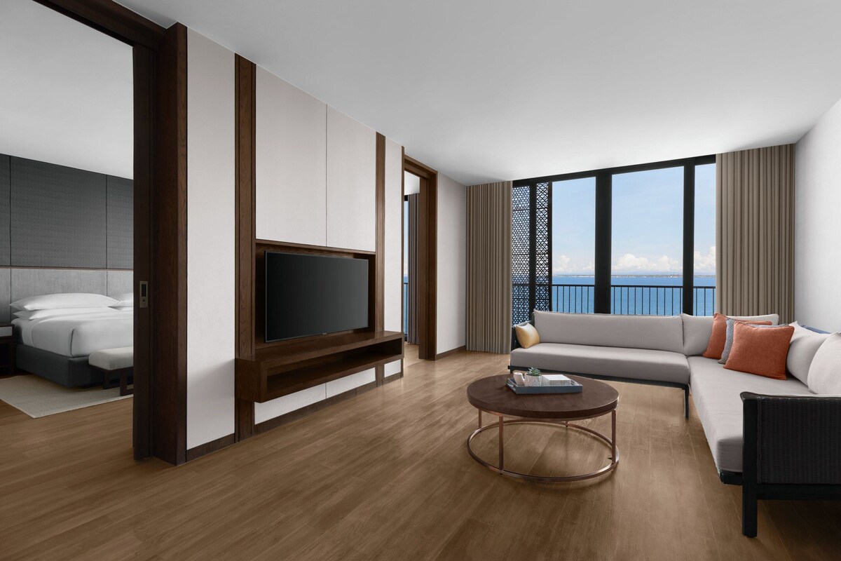 Habitación de hotel moderna con televisor grande, sofá, mesa de centro y balcón con vistas.