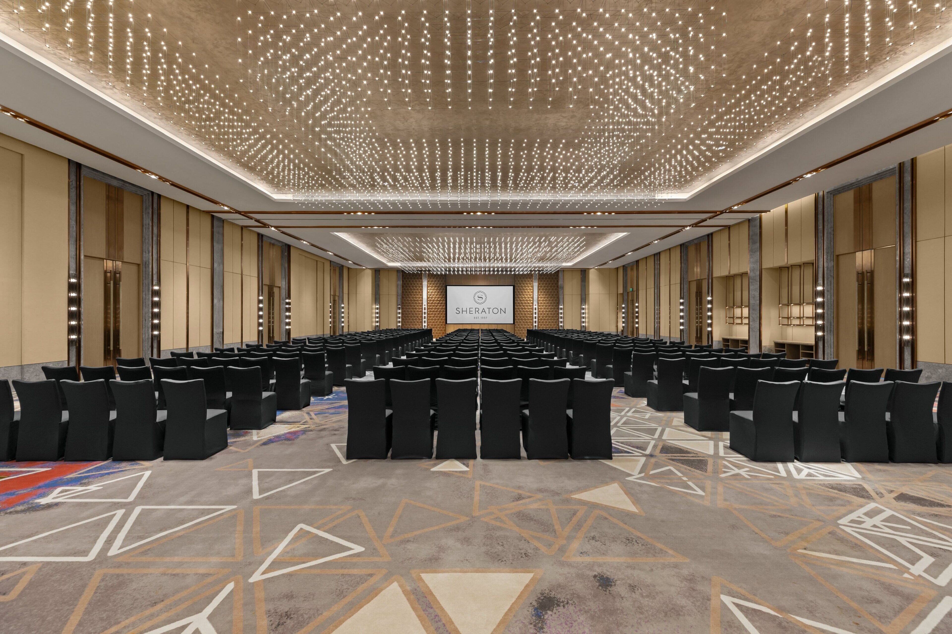 Une salle de conférence avec des rangées de chaises noires, une moquette à motifs et un plafond orné de luminaires.