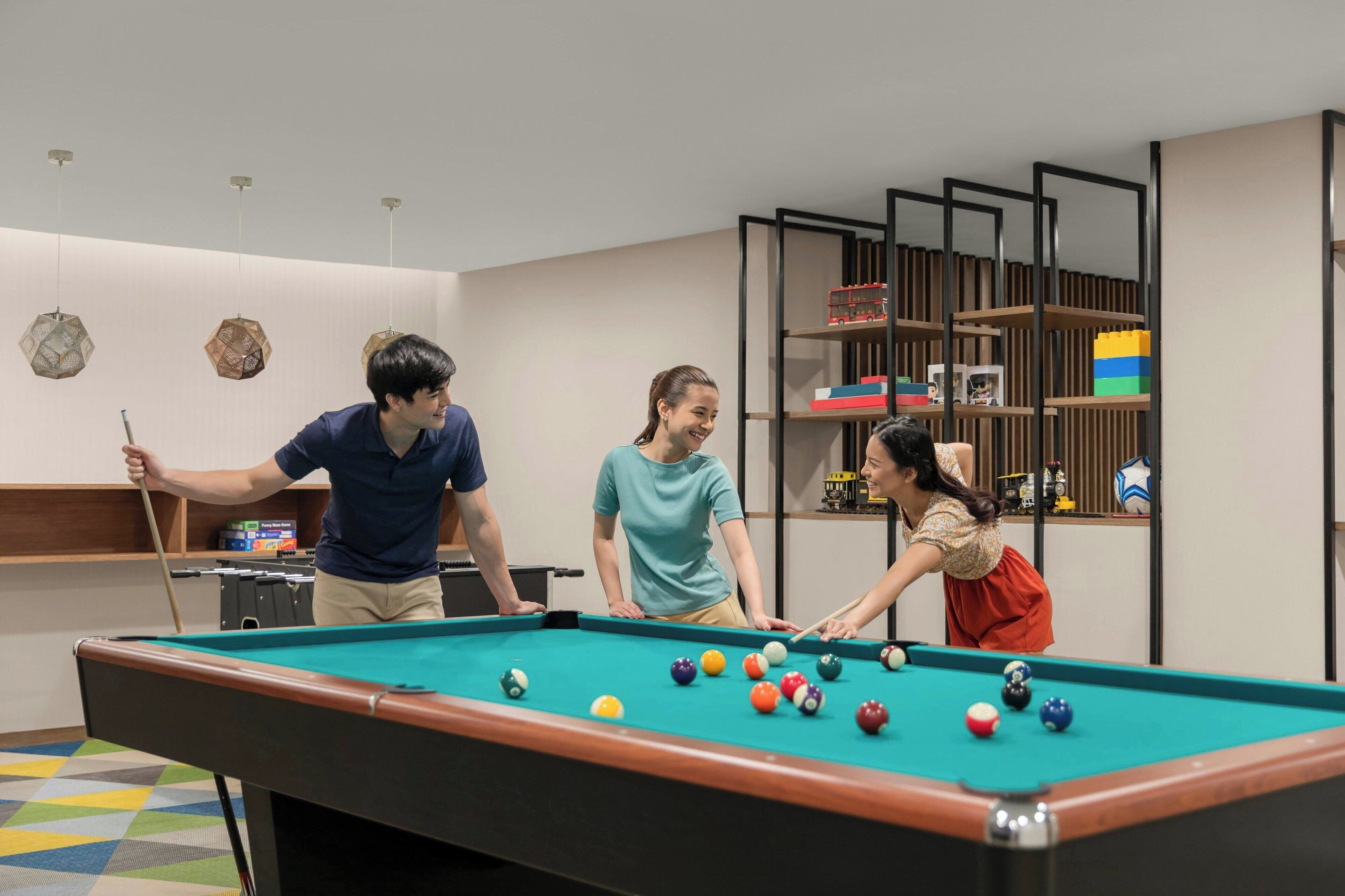Una mesa de billar con bolas, una persona sosteniendo una cuesa y un estante con varios objetos en una habitación con piso de diseño geométrico.