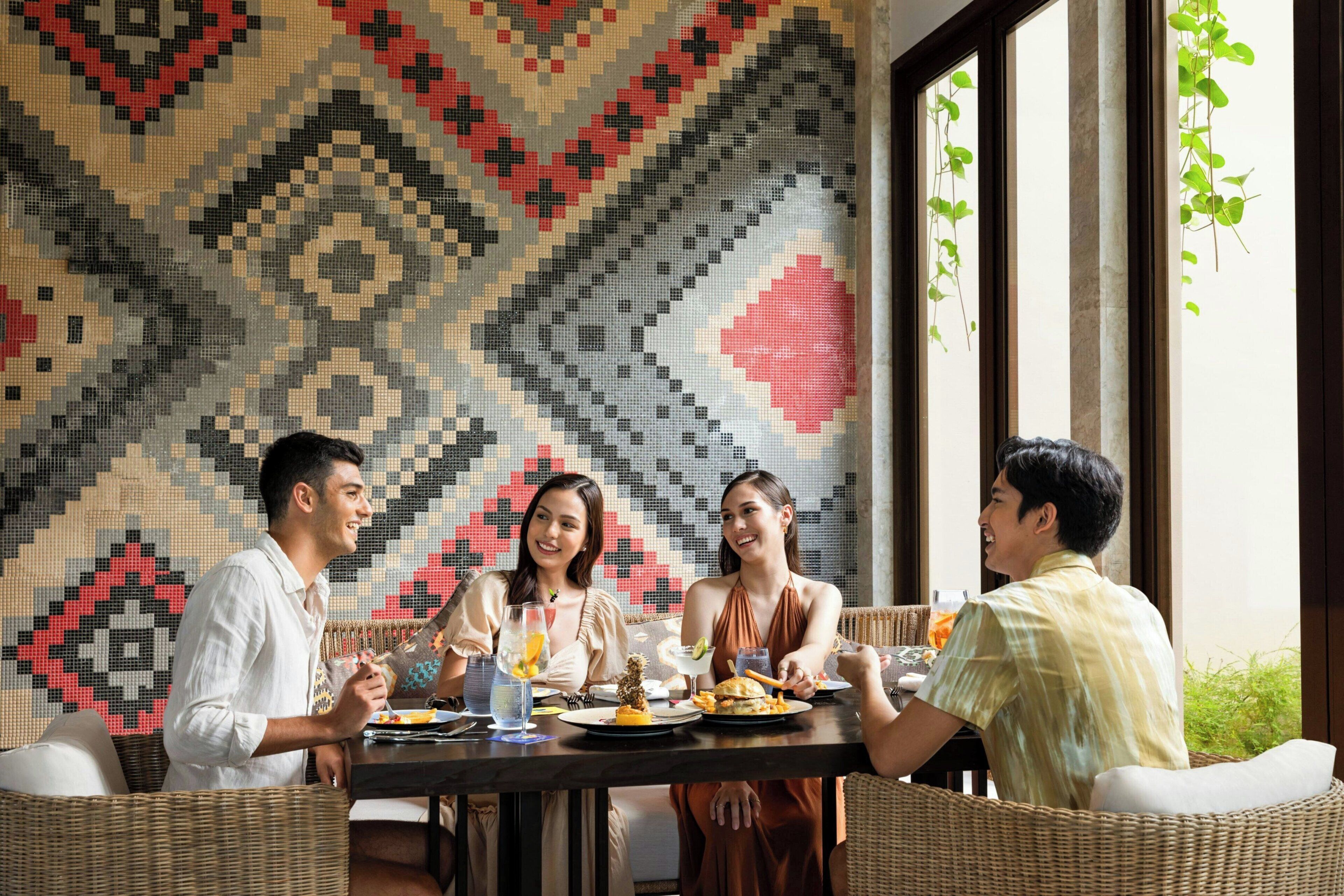 A group of people dining at a table with a patterned wall and large windows in the background.