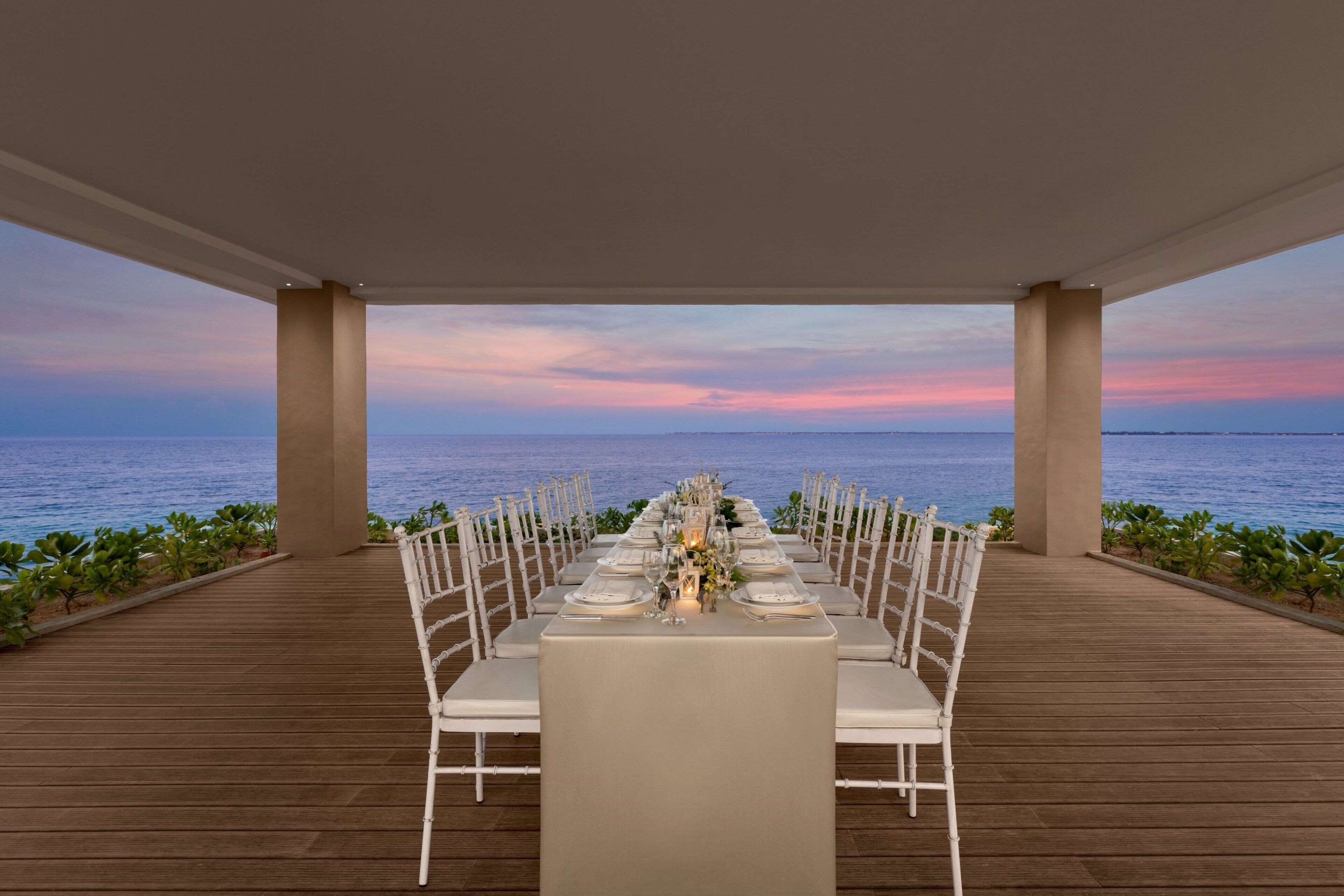 A long dining table set for a meal with a view of the sea at sunset.