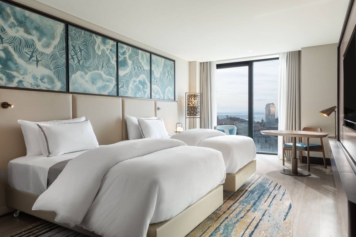 Une chambre d’hôtel avec deux lits, une grande fenêtre donnant sur la ville, une petite table avec une chaise et un élément décoratif mural.