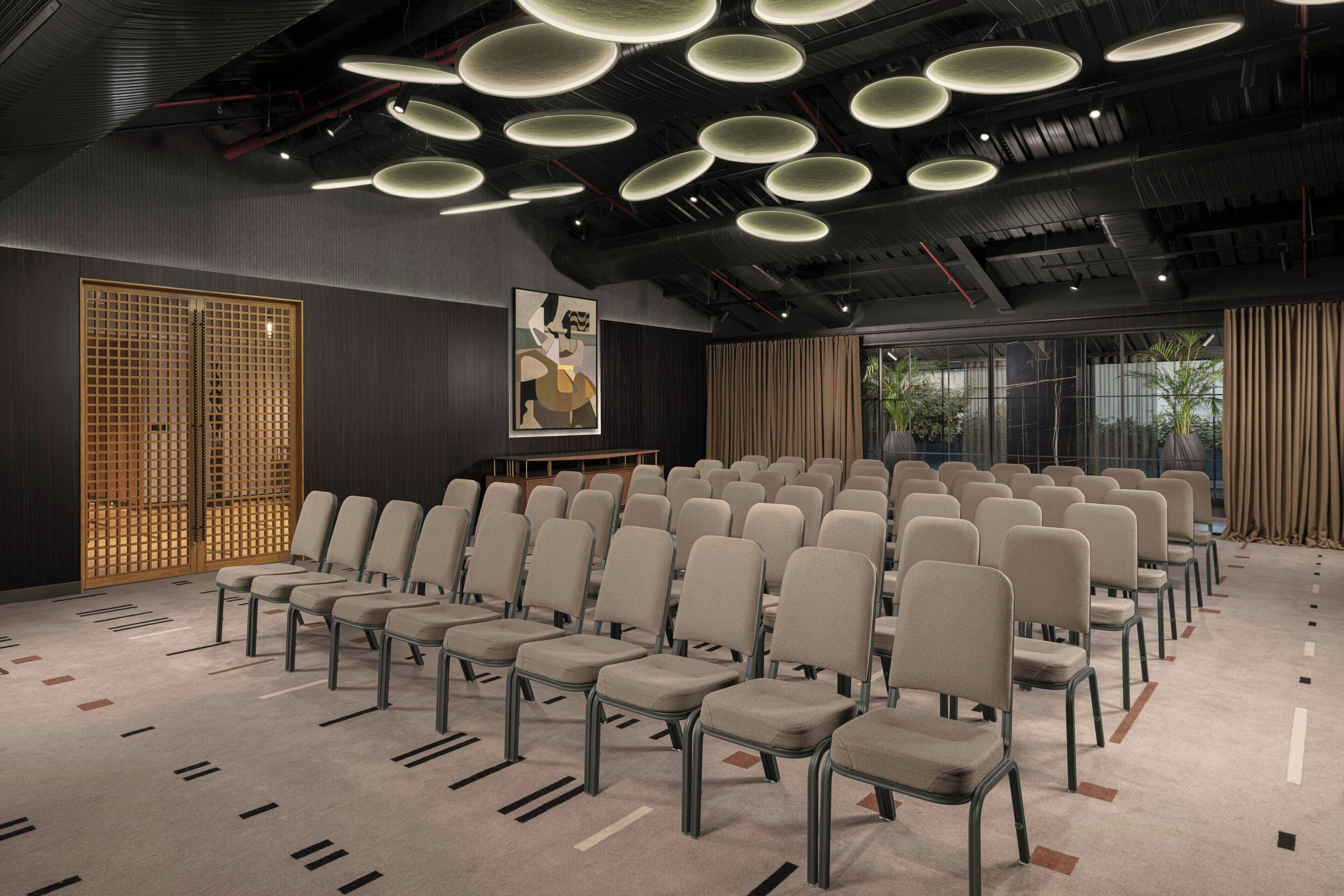 Een vergaderzaal met rijen stoelen, een groot raam met gordijnen en een plafond met ronde verlichtingsarmaturen.