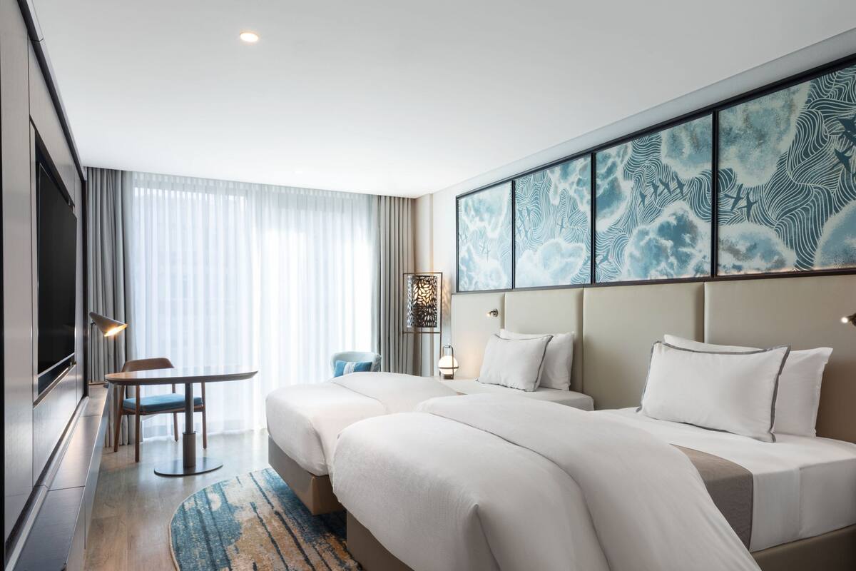 Une chambre d’hôtel avec deux lits, un bureau, une chaise, une télévision et une grande œuvre d’art accrochée au mur.