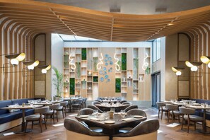 Een modern restaurantinterieur met meerdere eettafels en zitjes, een golvend plafond en een centrale wand met decoratieve borden en groen.