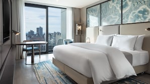 Une chambre d’hôtel moderne dotée d’un grand lit, d’une table à manger et offrant une vue sur le paysage urbain grâce à de grandes fenêtres du sol au plafond.