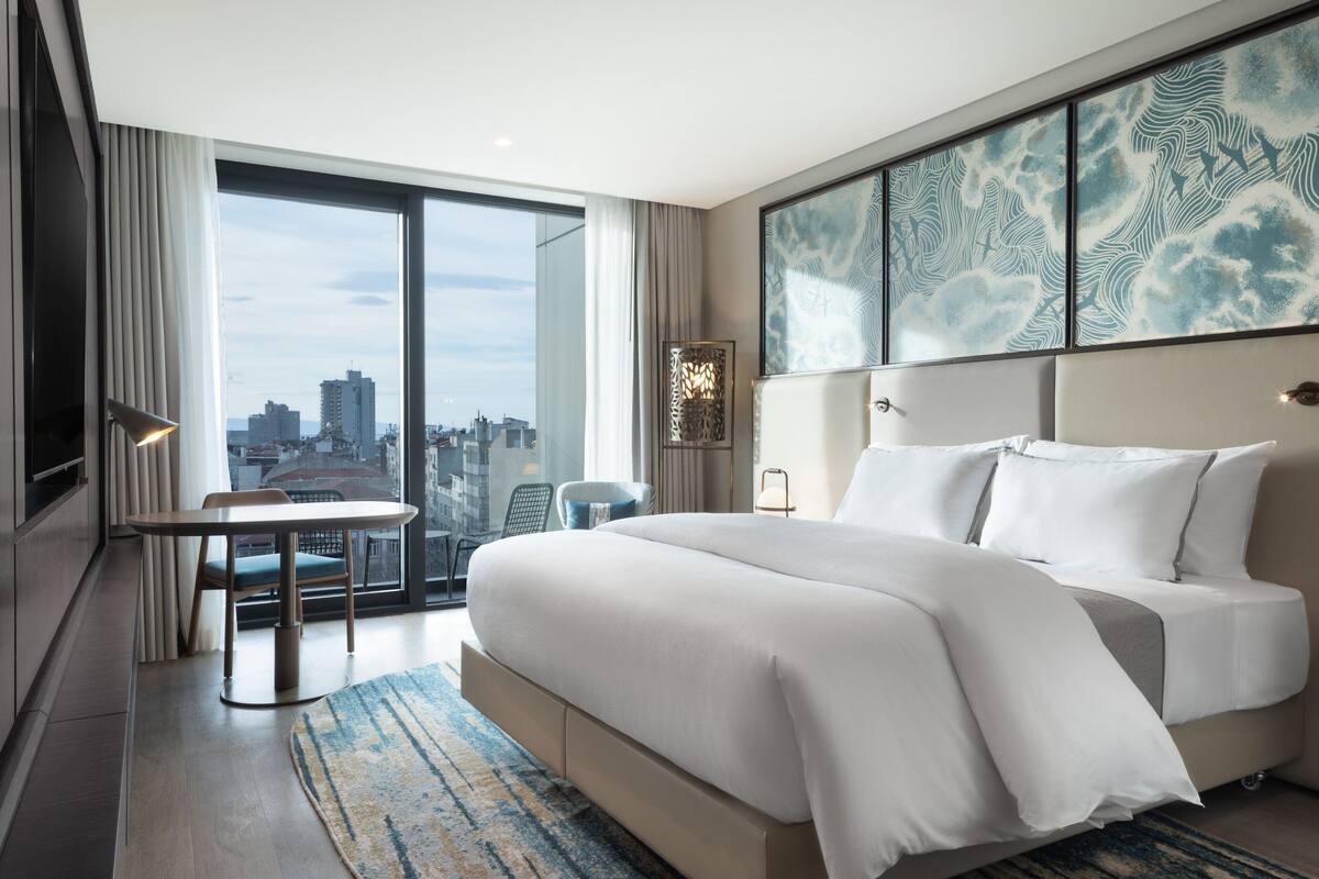 Une chambre d’hôtel moderne dotée d’un grand lit, d’une table à manger et offrant une vue sur le paysage urbain grâce à de grandes fenêtres du sol au plafond.