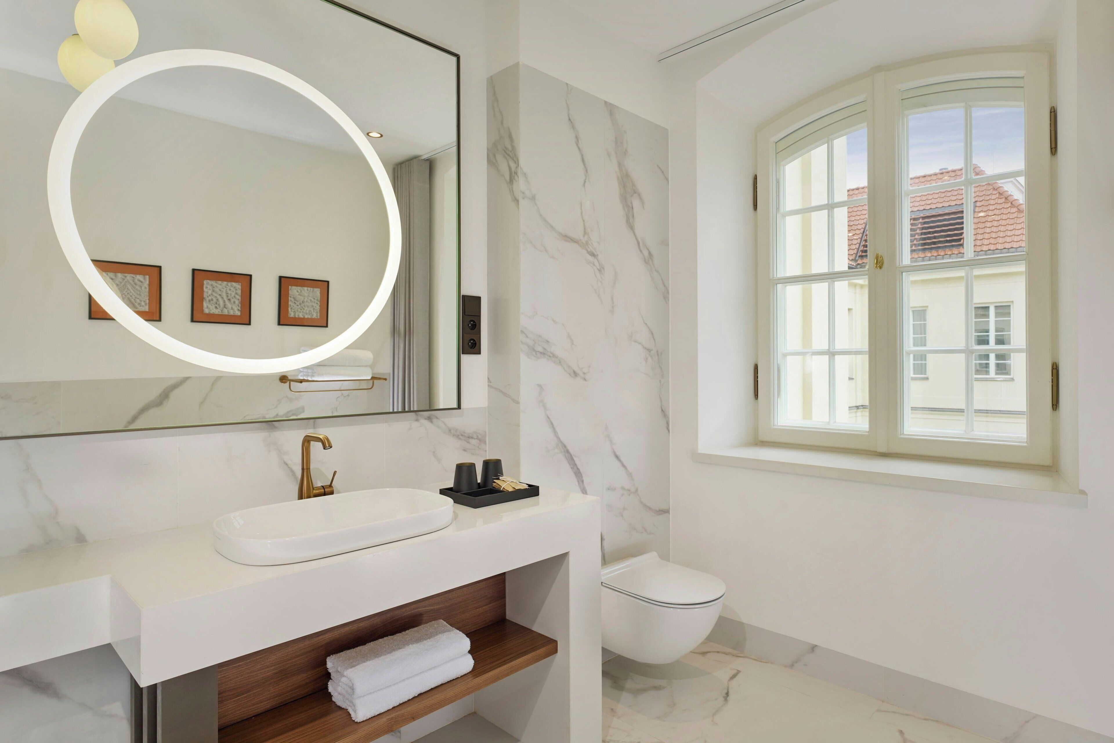 A modern bathroom with a white sink, a mirror with a circular light, a toilet, and a window with a view of buildings.