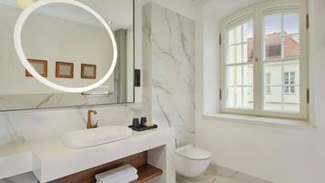 A modern bathroom with a white sink, a mirror with a circular light, a toilet, and a window with a view of buildings.