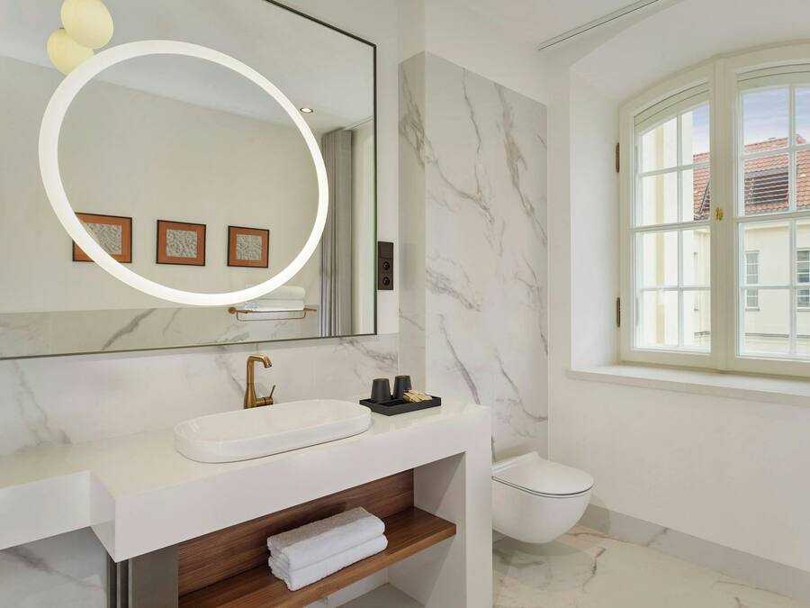 A modern bathroom with a white sink, a mirror with a circular light, a toilet, and a window with a view of buildings.
