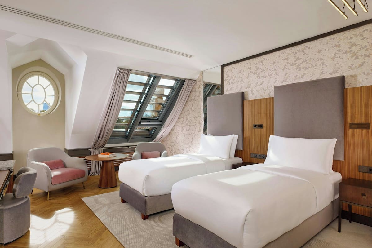 A hotel room with two beds, a sitting area with a chair, a small table, and a large window with curtains.