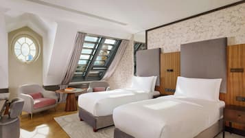 Habitación de hotel con dos camas, un rincón de descanso con una silla, una mesita y un amplio ventanal con cortinas.