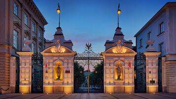Una entrada imponente con puertas decoradas y estatuas, flanqueada por edificios de estilo clásico.