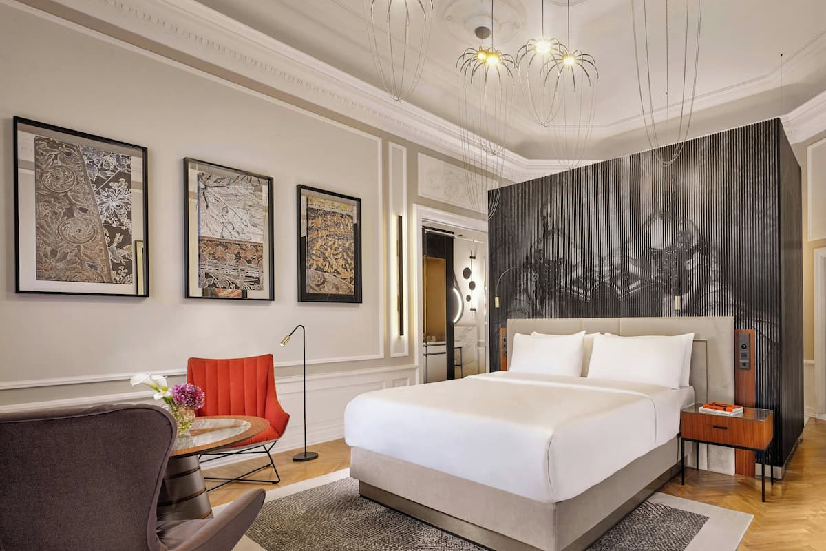 A modern hotel room with a large bed, a red chair, a small round table with a vase of flowers, and artwork on the walls.