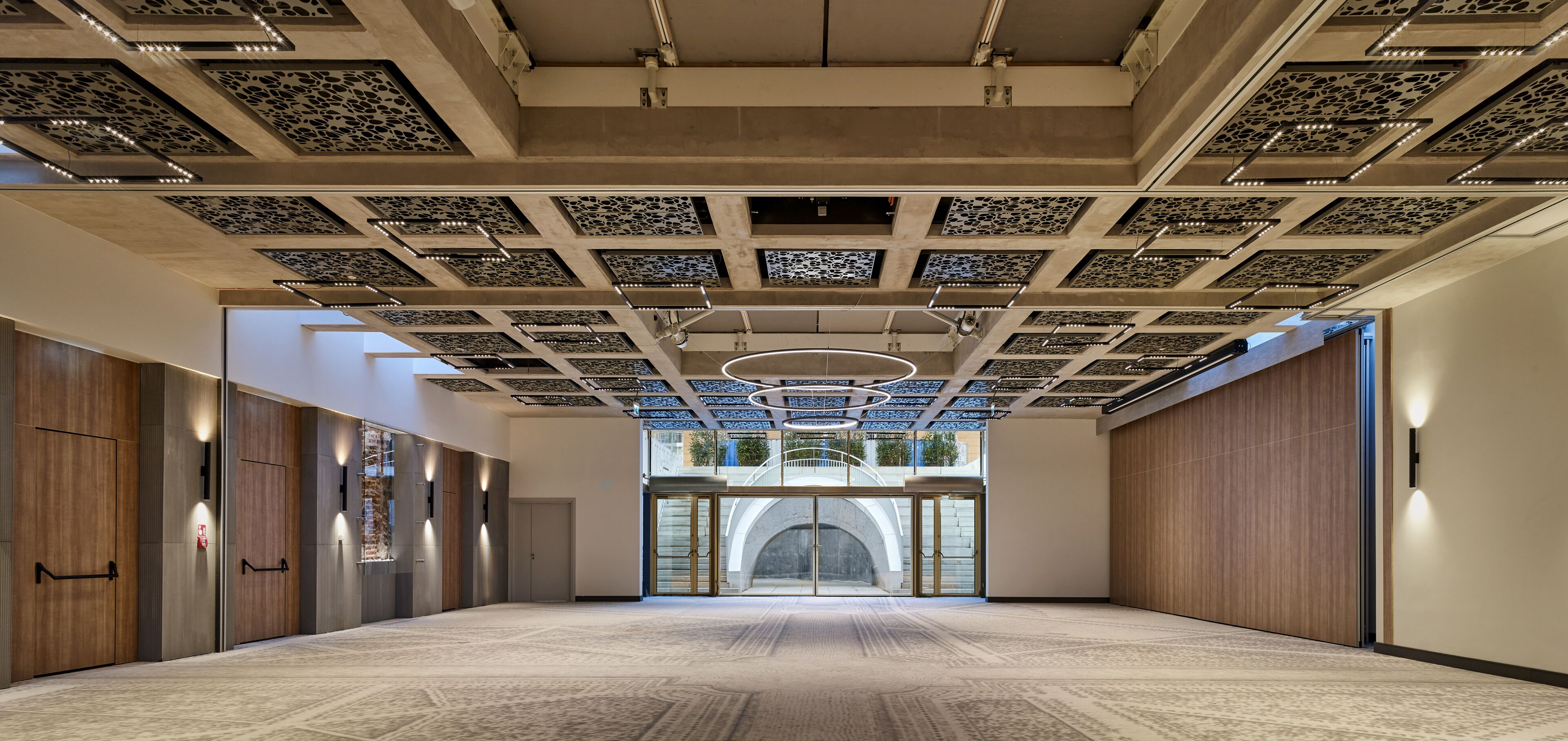 A spacious hallway with a patterned ceiling, wooden doors, and a view of an outdoor area.