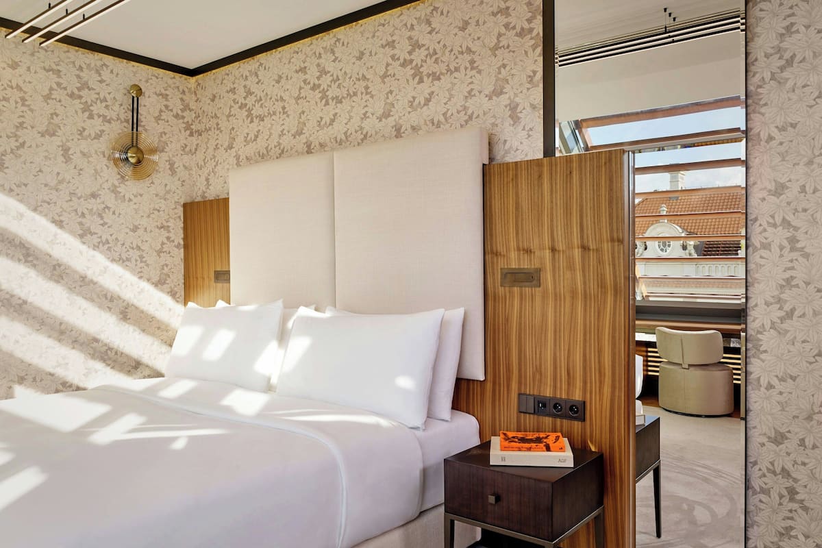 A modern hotel room with a large bed, a wooden headboard, a nightstand with a book, and a view of the outside.