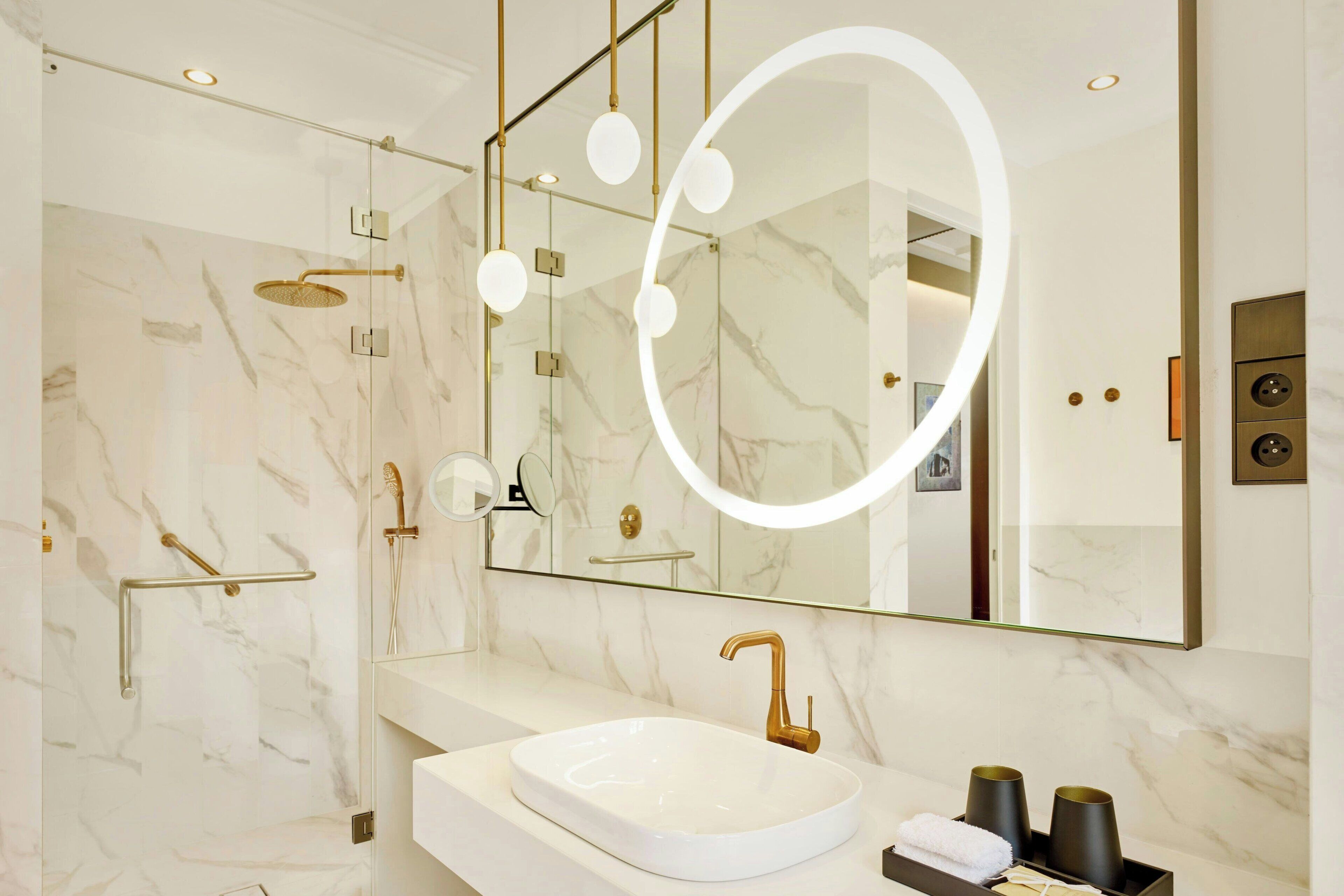 Un baño moderno con lavamanos de mármol, elementos dorados y un espejo grande con luz circular.