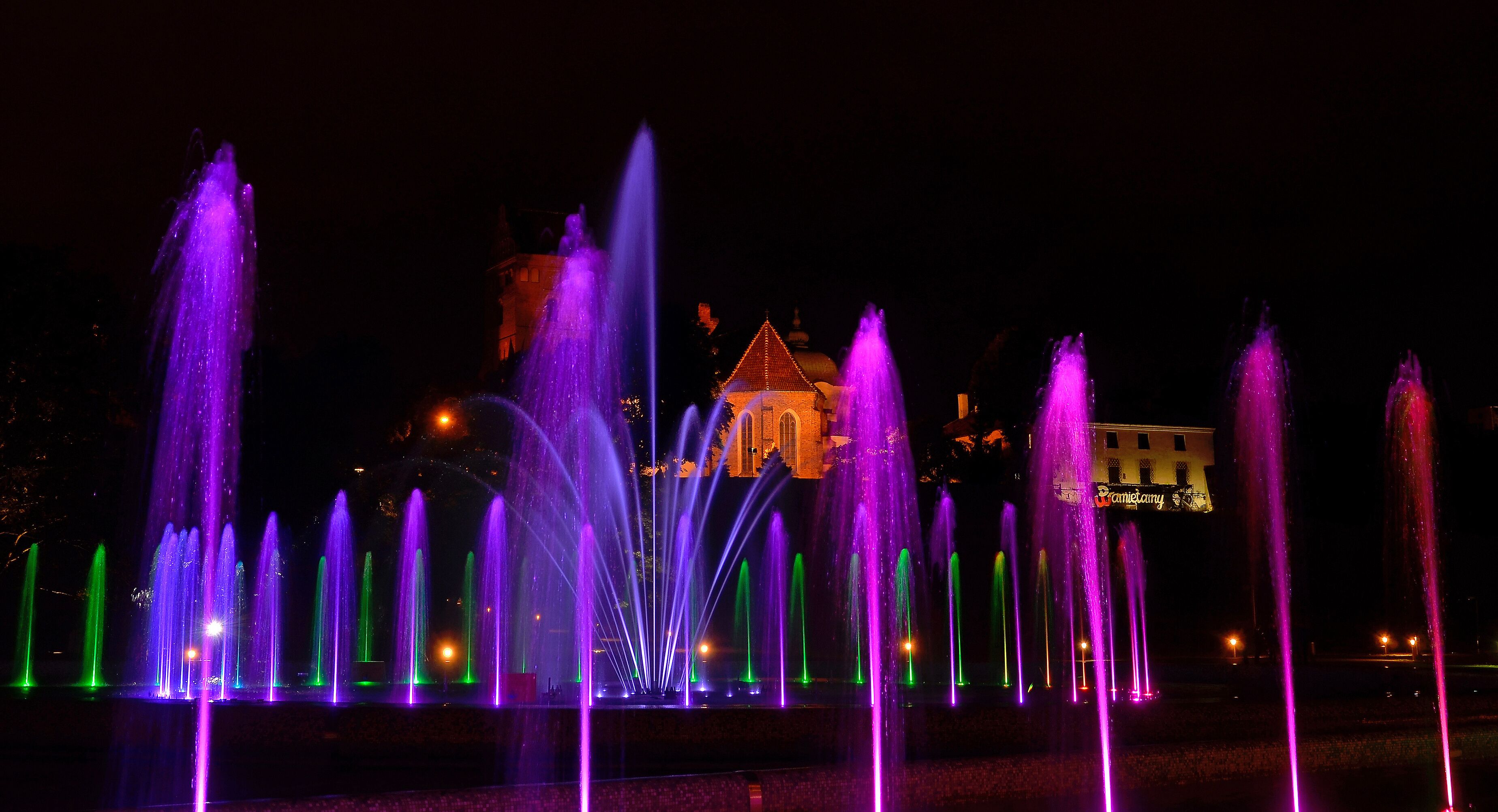 Vista nocturna de una fuente con múltiples chorros de agua iluminados con luces moradas y verdes, y un edificio al fondo.