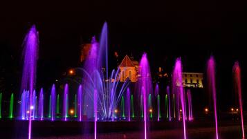 Vista nocturna de una fuente con múltiples chorros de agua iluminados con luces moradas y verdes, y un edificio al fondo.