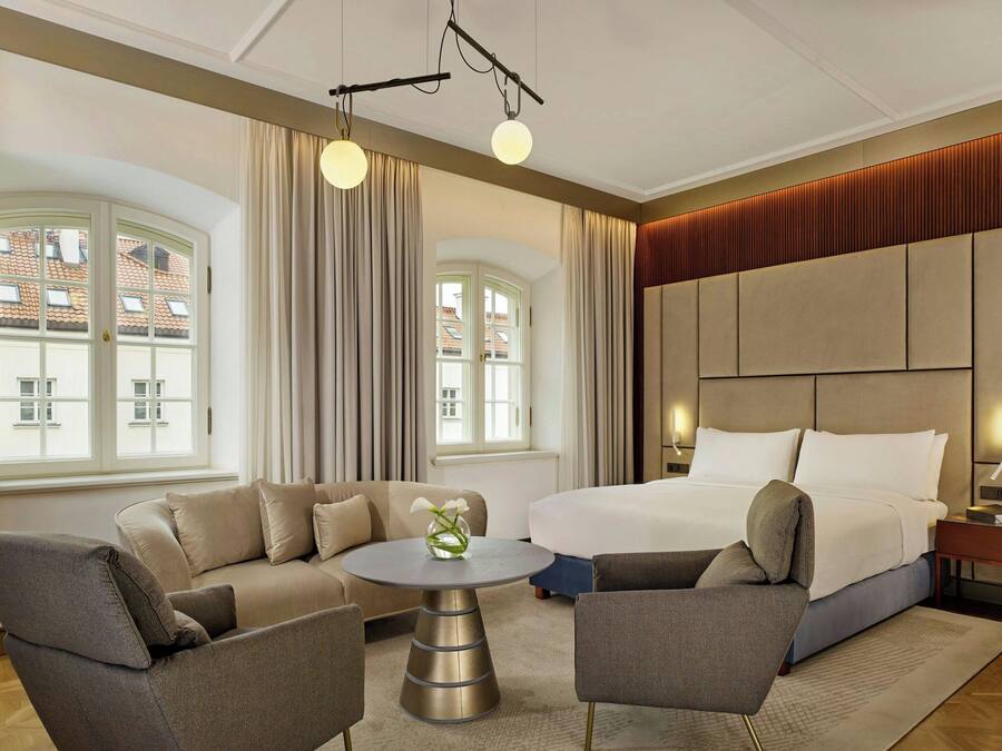 A modern hotel room with a large bed, two armchairs, a round table, and a view of buildings through large windows.