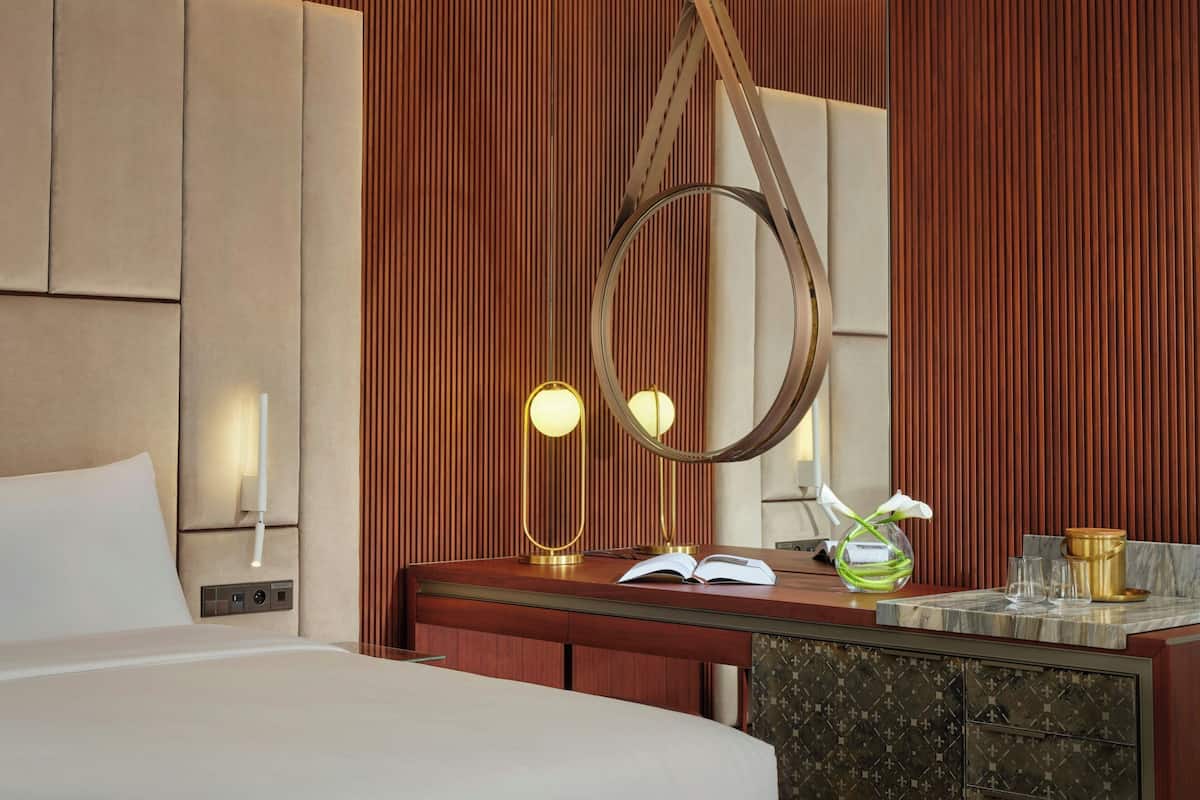 A hotel room with a bed, a wooden headboard, a bedside table with a lamp, a mirror, and a vase with flowers.