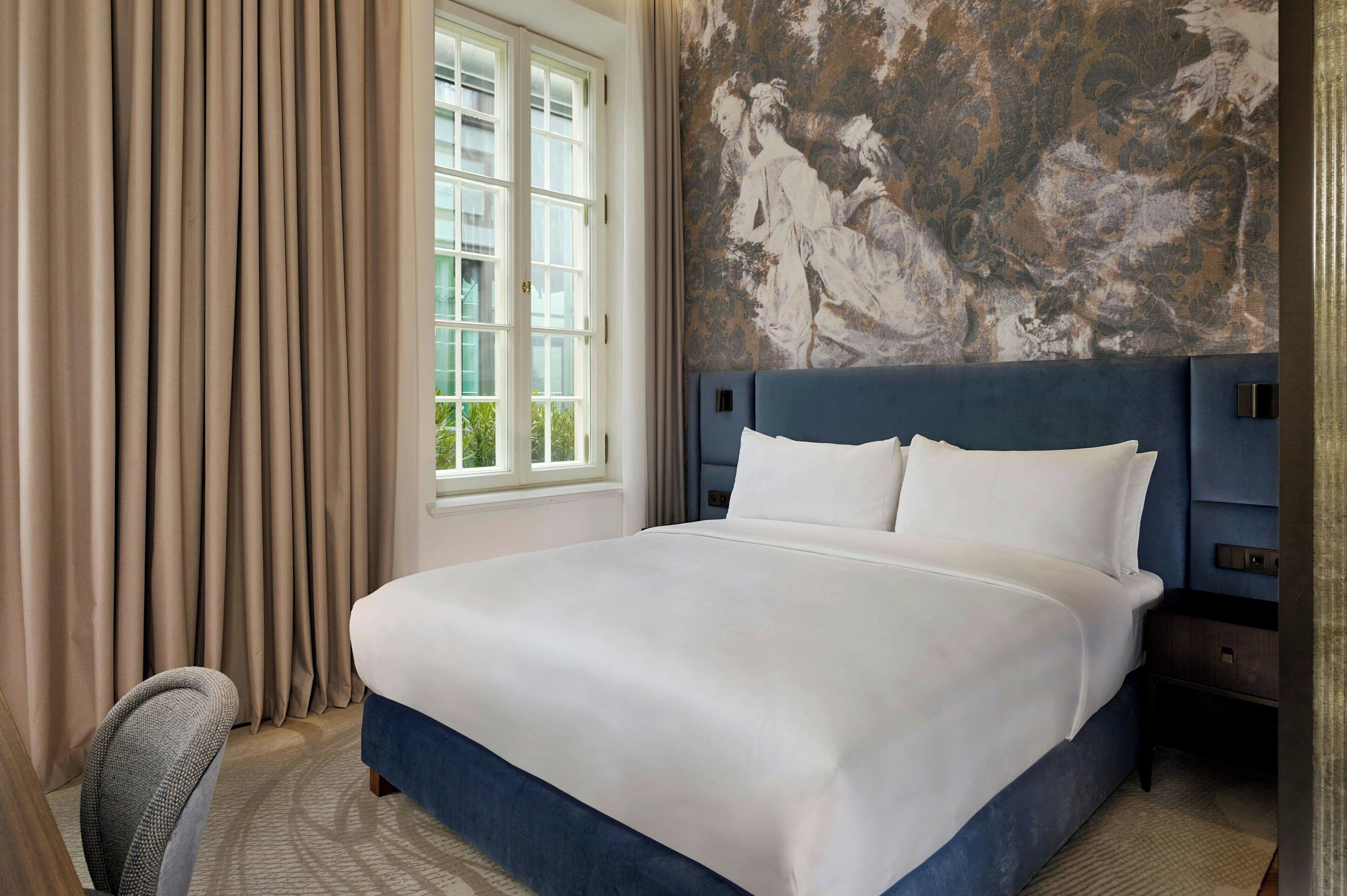 Ein Hotelzimmer mit einem großen Bett, einem Kopfteil, einem Fenster mit Vorhängen und einer Wand mit Wandgemälde.