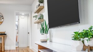 Ein modernes Wohnzimmer mit einem wandmontierten Flachbildfernseher, Holzregalen mit Pflanzen, einem weißen Konsolentisch und Holzböden.