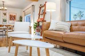 A living room with a brown leather sofa, white armchairs, and a small white table. There is a ladder with books and a lamp against the wall.