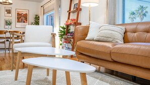 Ein modernes Wohnzimmer mit einem braunen Ledersofa, weißen Sessel und einem weißen Couchtisch. Eine Leiterregalwand beherbergt Bücher und Dekorationsobjekte, und ein großes Fenster mit Vorhängen.