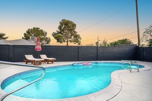 A swimming pool with lounge chairs and inflatable rings, surrounded by a dark wall and trees. - Modern • Tesla charger • 10 mins from Strip+Airport (Las Vegas)