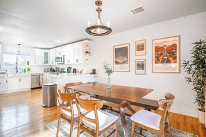 A modern kitchen with white cabinets, a wooden dining table, and chairs. There are framed pictures on the wall and a plant on the right. - Modern • Tesla charger • 10 mins from Strip+Airport (Las Vegas)
