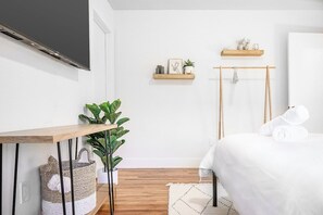 A bedroom with a bed, a wooden bedside table, a wall-mounted TV, a plant, a hanging towel rack, and a small shelf with decorative items.
