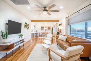 A modern living room with a flat-screen TV, a ceiling fan, a brown leather sofa, and a dining area in the background. - Modern • Tesla charger • 10 mins from Strip+Airport (Las Vegas)