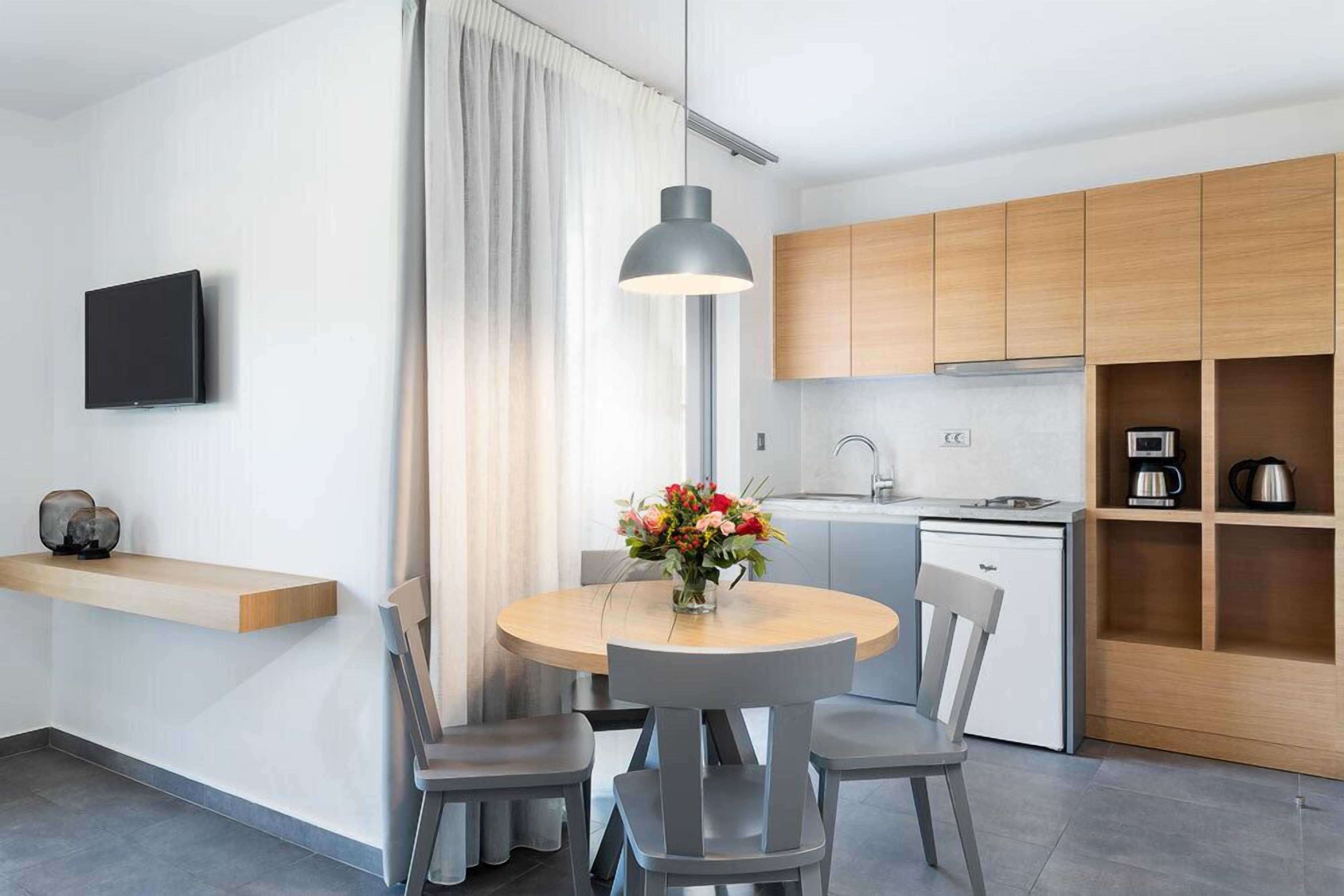 A modern kitchen with wooden cabinets, a dining table with four chairs, a vase of flowers, and a flat-screen TV on the wall.