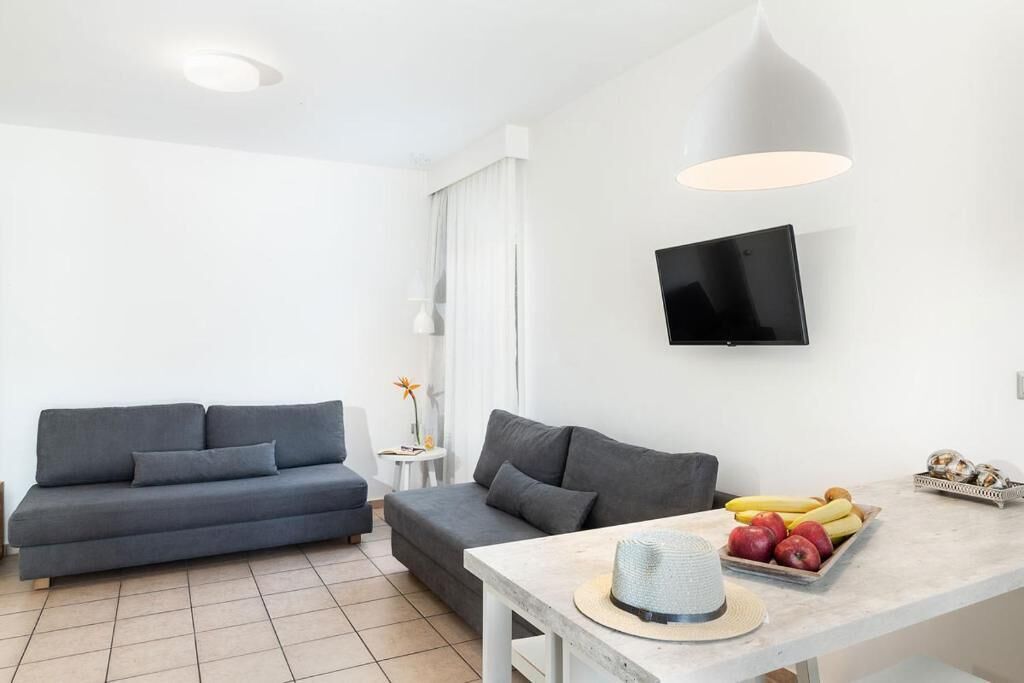 A modern living room with a grey sofa, a small round table with fruits, and a TV mounted on the wall.