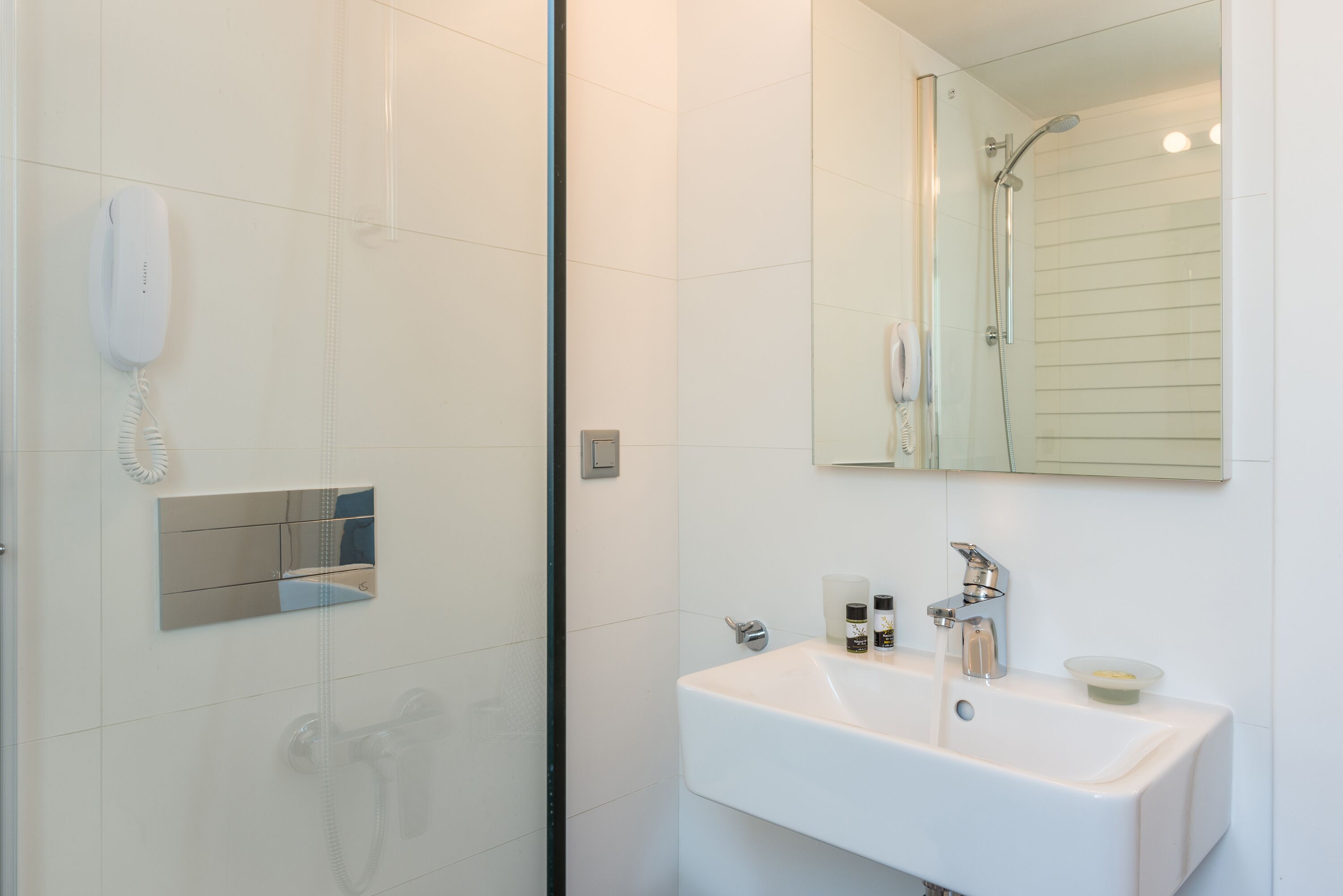 Un baño moderno con lavamanos blanco, un espejo, un cabezal de ducha manual y un teléfono.