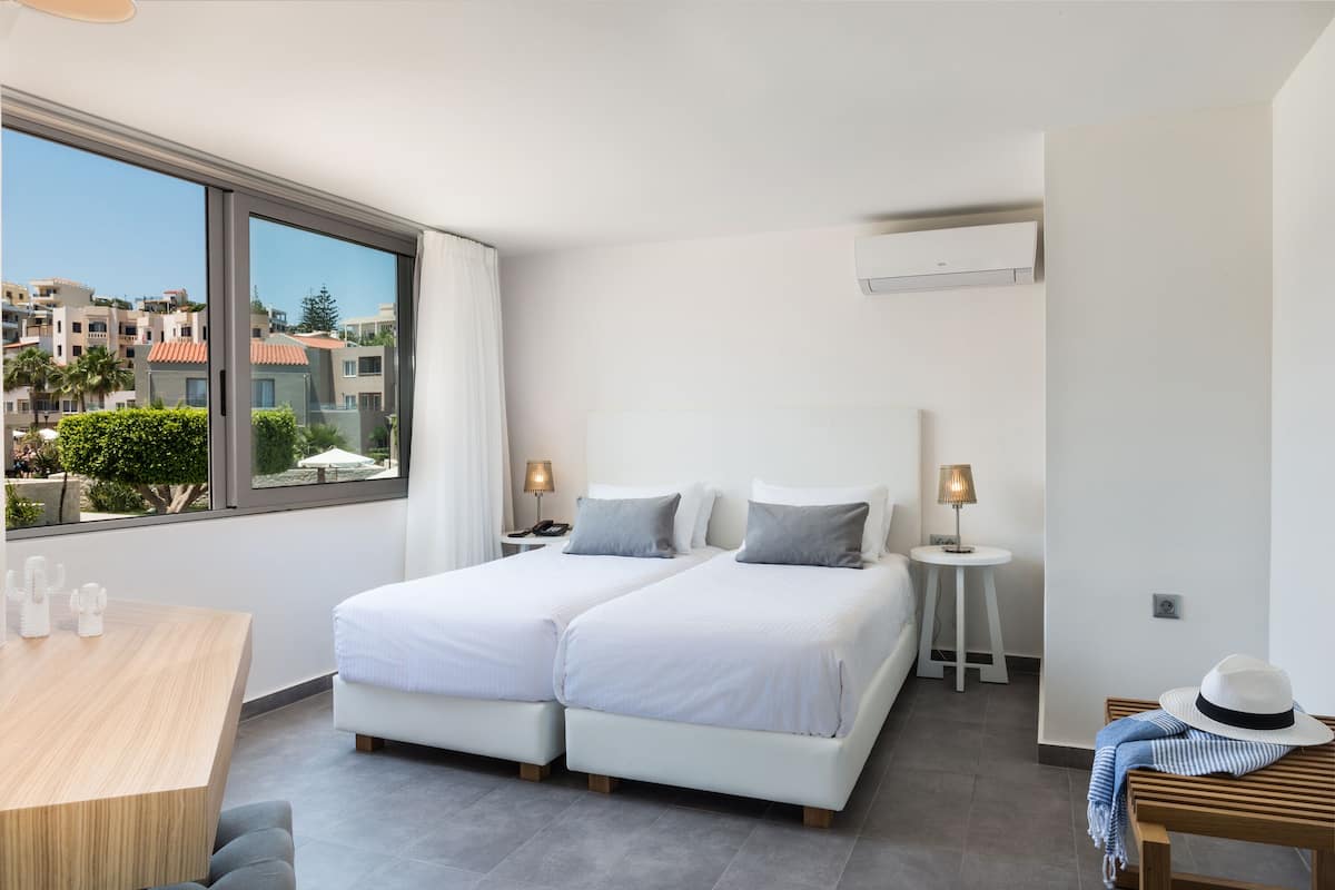 A modern hotel room with two beds, a wooden table, and a view of the cityscape.