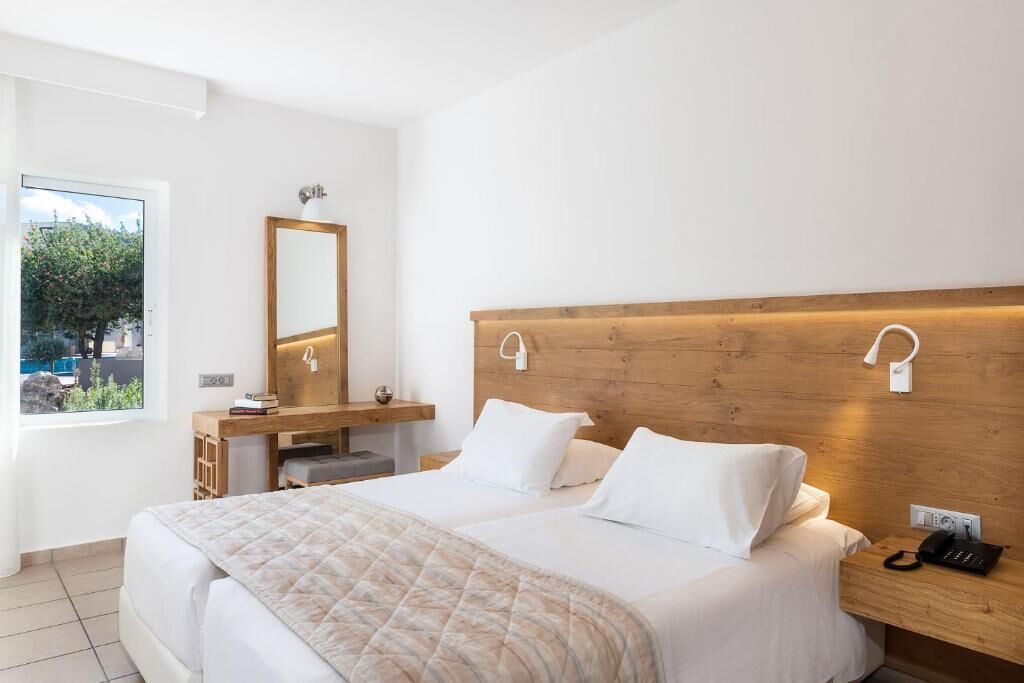 A modern hotel room with a large bed, a wooden headboard, a bedside table with a phone, a mirror, and a view of the outdoors through a window.