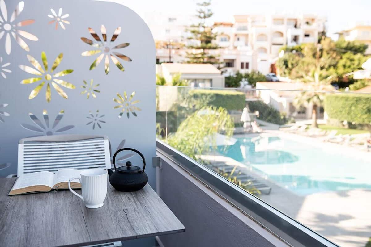 A balcony with a view of a swimming pool, a white chair, a table with a book and a white mug, and decorative wall art.